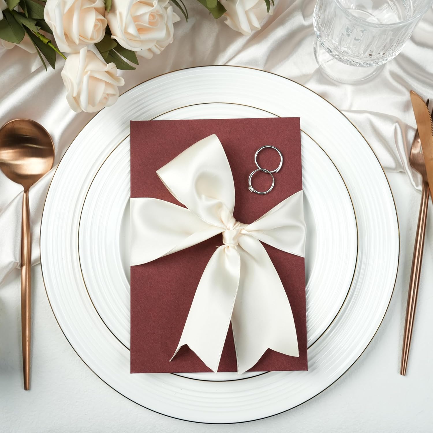 Invitație de nuntă burgundy decorată cu panglică satin ivory, aranjată pe farfurie albă cu tacâmuri aurii și trandafiri crem – Diandra Events