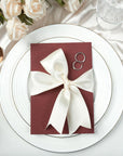 Invitație de nuntă burgundy decorată cu panglică satin ivory, aranjată pe farfurie albă cu tacâmuri aurii și trandafiri crem – Diandra Events