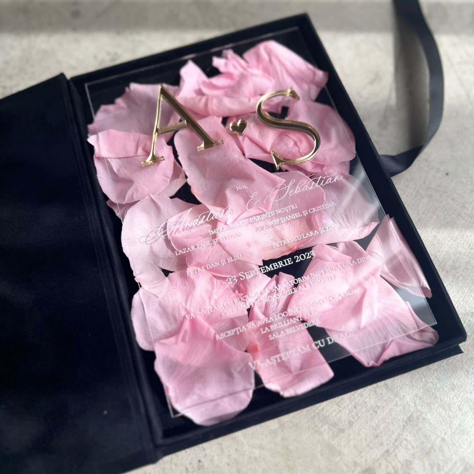 Invitație de nuntă acrilică transparentă cu inițiale aurii și petale roz de trandafir, prezentată într-o cutie de catifea neagră