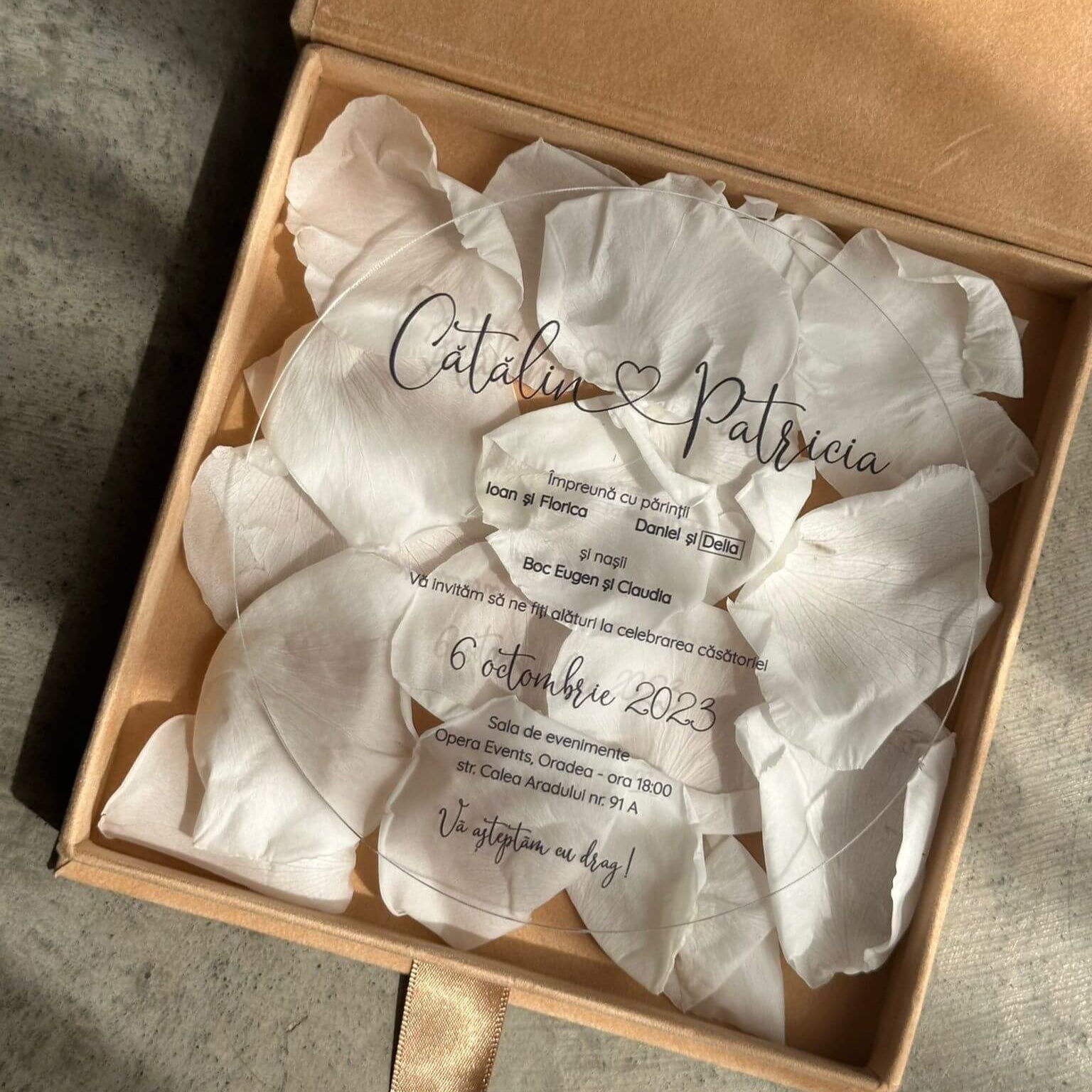 Invitație de nuntă din acril transparent, prezentată într-o cutie elegantă cu petale albe de trandafir