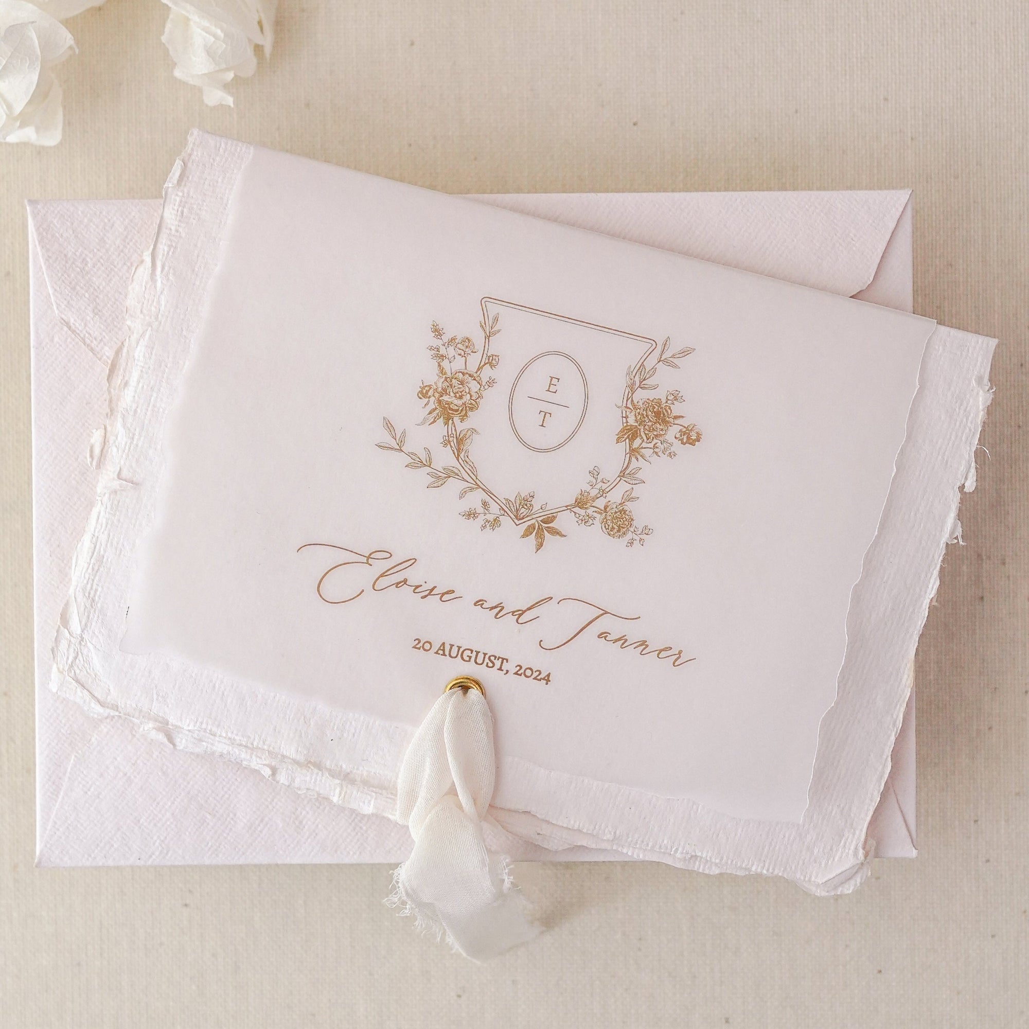 Invitație de nuntă cu monogramă florală aurie, printată pe hârtie handmade cu margini neregulate, învelită în vellum transparent și legată cu panglică din mătase – model elegant Diandra Events